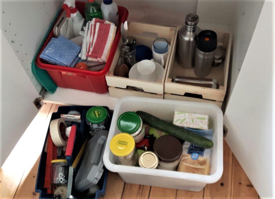 Blick in Küchenunterschrank: rote Kiste mit Putzuntensilien, 2 Holzkästen mit einigen Küchenutensilien. Vor dem Schrank stehend: 1 Werkzeugkiste, rechts daneben eine weiße Box mit Nahrungsmittelvorräten