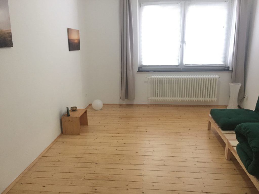 Zimmer mit Holzdielenboden, einem Hocker 2 Bildern. Am rechten Bildrand ist ansatzweise ein grünes Futon zu sehen