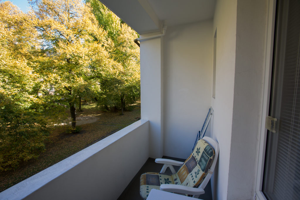 Überdachter Balkon mit Balkonstuhl und Blick in einen Innenhof mit großen Bäumen