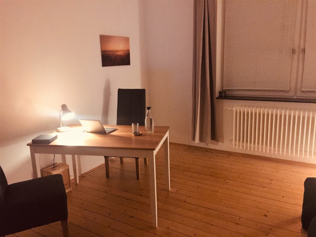 Foto mit abendlicher Stimmung: Tisch mit 2 Stühlen und Laptop und kleiner Lampe. Im Hintergrund ein Fenster. 