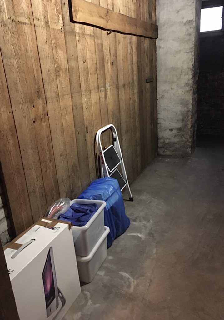 Kleiner Keller mit links an der Wand stehendem Karton, 2 Plastikkisten, ein blauer Müllsack und eine kleine Trittleiter