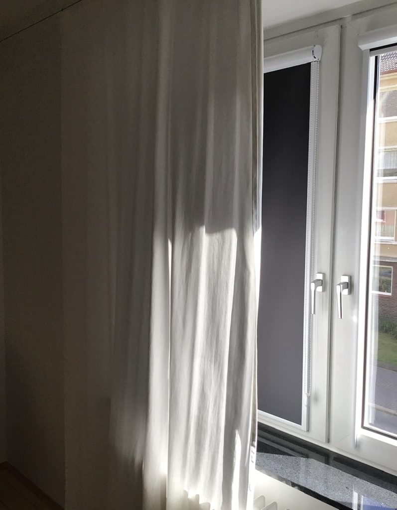 Blick auf ein Fenster mit herunter gelassenem Rolle und einem weißen Vorhang. Rechts ansatzweise ein geöffnetes Fenster, durch das Licht scheint.