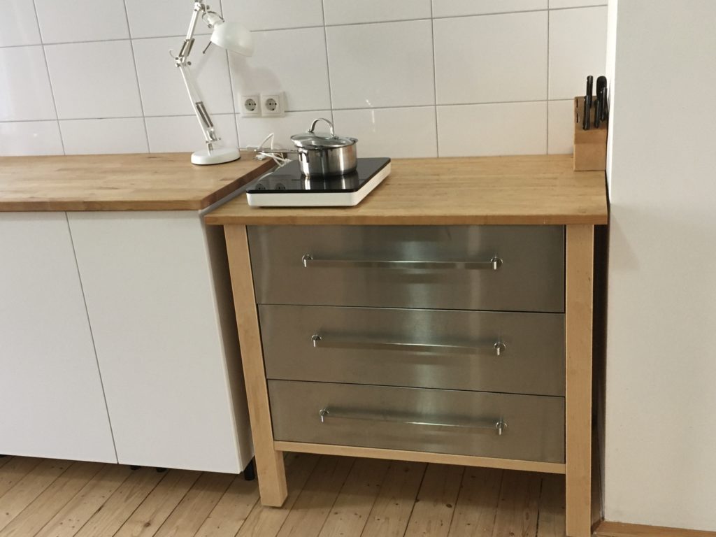 Schubladenschrank mit Kochplatte und einem Stieltopf darauf. Rechts ein Messerblock, links eine Schreibtischleuchte auf der Arbeitsplatte