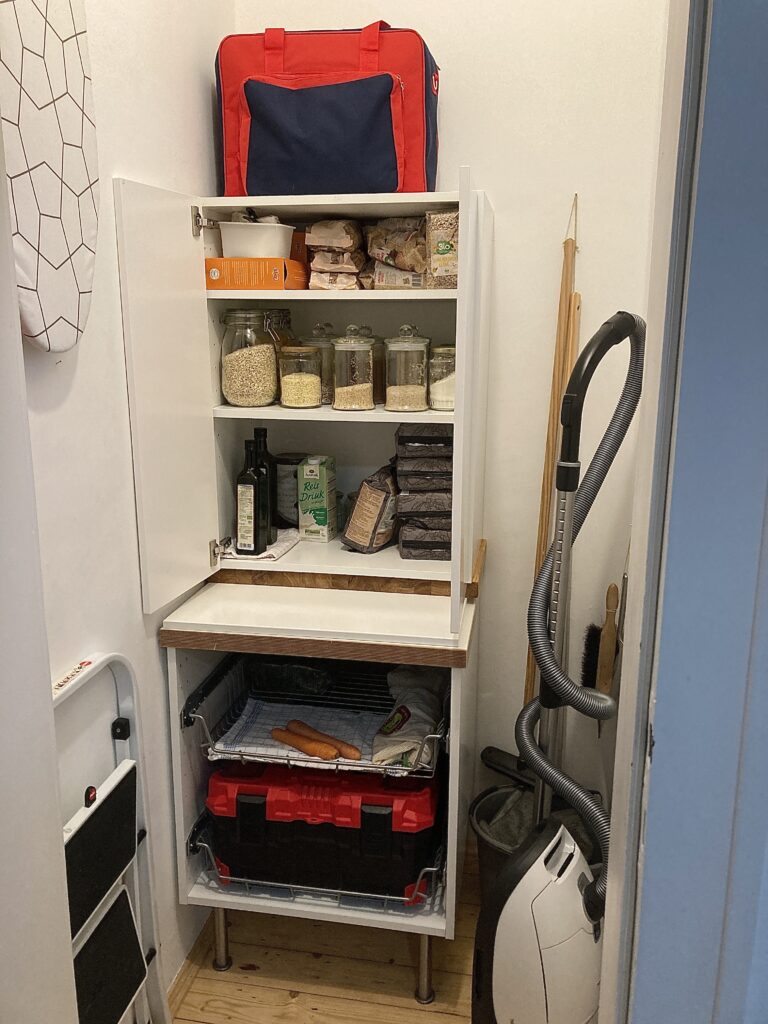 Abstellkammer mit Unterschrank und geöffnetem Oberschrank und diversen Küchenvorräten darin. Rechts daneben Staubsauger und einige Putzutensilien