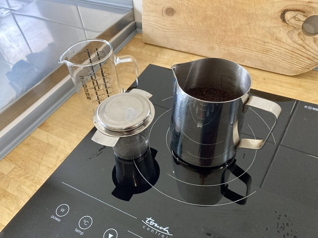 Kochplatte mit Edelstahl-Milchkännchen. Darin Kaffeepulver und Wasser. Links daneben ein Teesieb