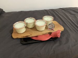 Joghurt-Milchmasse in Gläsern auf Holzbrett. Darunter Wärmflasche, die auf einem Backblech liegt. Alles auf einem Bett stehend.